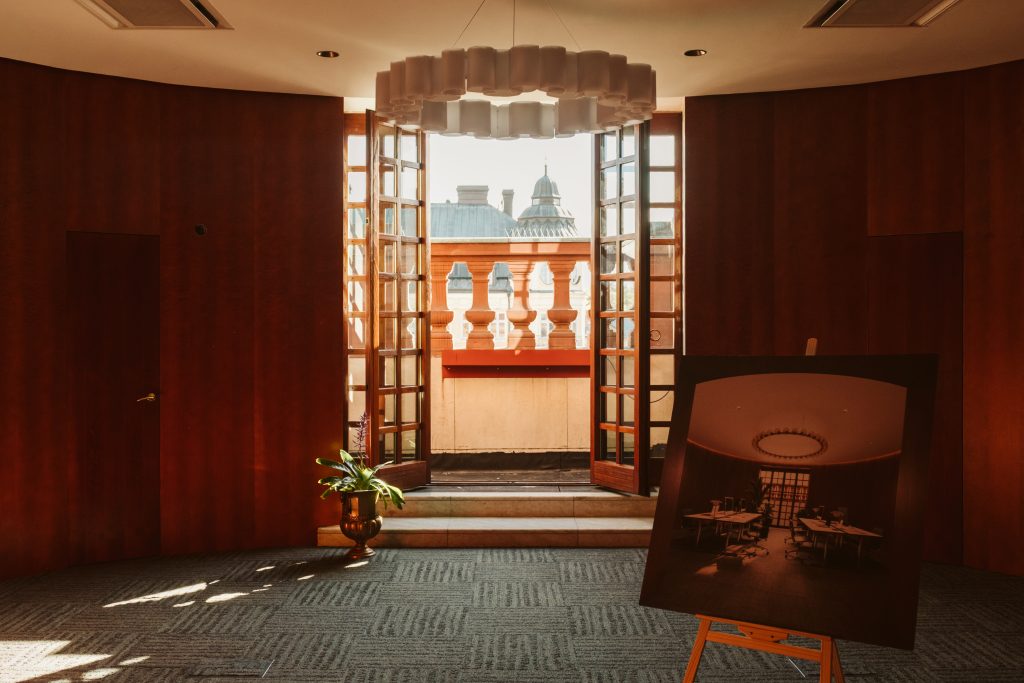 Interiör hos Banken med ljusa kontorsrum, stora fönster, trägolv och dekorativa träpaneler sedda genom en öppen dörr.