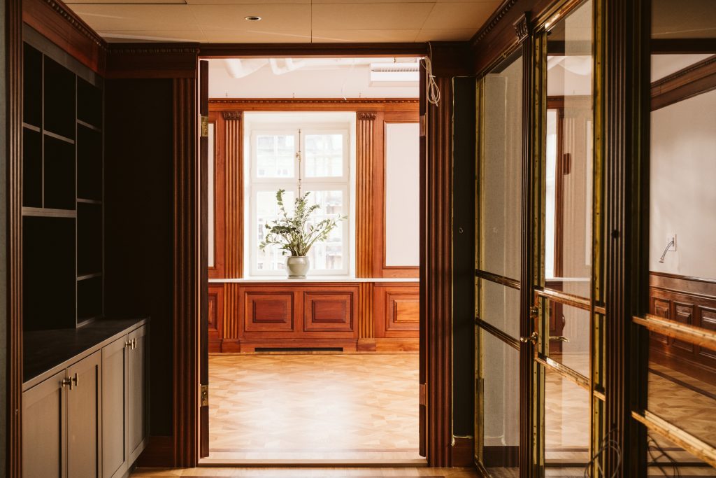Interiör hos Banken med ljusa kontorsrum, trägolv, stora fönster och dekorativa träpaneler sedda genom en öppen dörr.